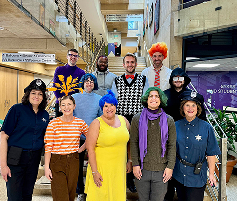 A group of staff members pose together inside the Sykes Student Union, wearing colorful costumes for a Halloween event. The group stands on and around a staircase in the building’s atrium, with individuals dressed in a variety of themed outfits, including wigs, hats, uniforms, and bright clothing. Directional signage, railings, and the open interior of the student union are visible in the background.