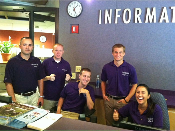 A group of five student staff members pose behind an information desk inside a campus building. They wear matching purple polo shirts with embroidered text, stand or sit around the desk, and smile toward the camera, with one person giving a thumbs-up. The desk area includes pamphlets, a computer monitor, and a countertop, and a wall behind them displays large lettering that reads “Information,” along with a wall clock and office fixtures.