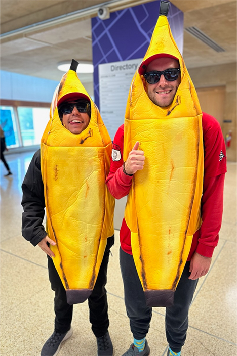 Two people standing indoors wearing full-body banana costumes. Both wear sunglasses and smile toward the camera, with one giving a thumbs-up.