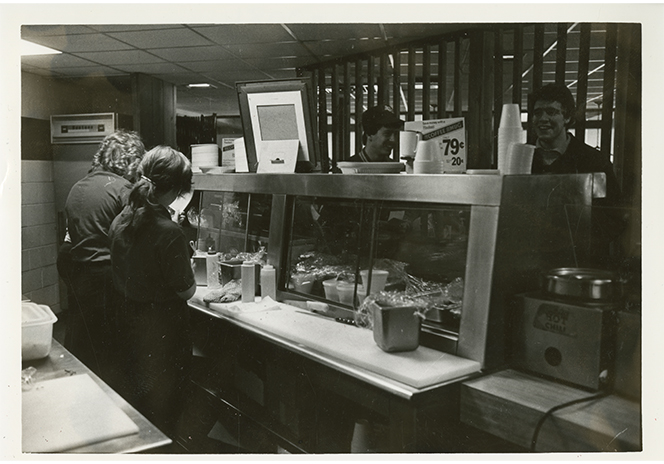 Black-and-white photograph of students ordering food at a deli counter inside a college union. Several students stand on one side of a glass display case while staff members work behind the counter preparing food. Condiment bottles, cups, and food containers line the counter, and a small price sign is visible above the serving area.