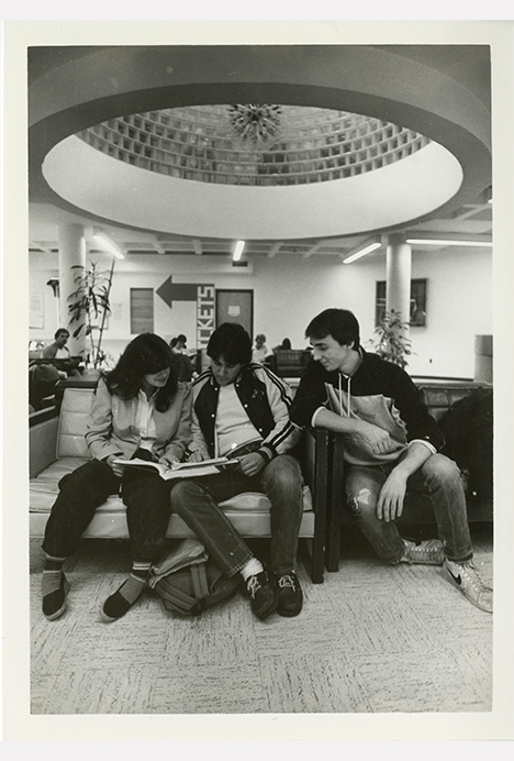 Black-and-white photograph of three students sitting together on a couch in the Sykes college union lobby. Two students in the center look down at an open book or notebook, appearing to study or read together, while the student on the right leans in and looks on. The space features cushioned seating, potted plants, and a large circular ceiling opening with a decorative light fixture above.
