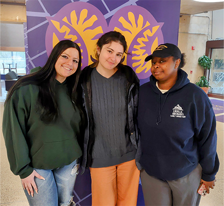 Three people standing together indoors and smiling at the camera. They pose in front of a purple pillar with a decorative painting of the West Chester University Logo in yellow. The individuals wear casual clothing, including hoodies and sweaters.