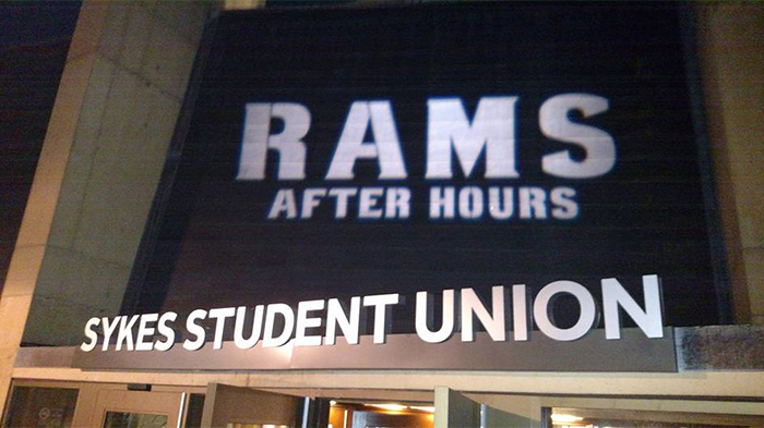 A nighttime photograph of the exterior entrance to the Sykes Student Union. Large spotlightted text on the building reads “RAMS After Hours,” with “Sykes Student Union” displayed below. The image shows the building facade lit from below, emphasizing the signage against the dark background.