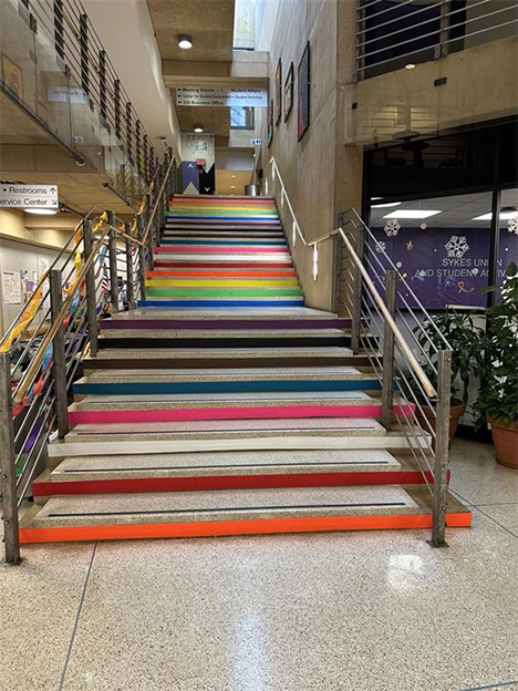 The main staircase inside the Sykes Student Union decorated for Love-is-Love Week. Each step is lined with bright colors from different identity flags, creating a rainbow effect up the stairs. Metal railings line both sides of the staircase, and directional signage and student union wall graphics are visible in the surrounding atrium space.