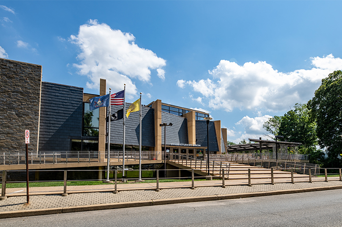 Exterior of the Sykes Student Union on a clear day. The modern building features dark paneling, stone accents, large windows, and an accessible ramp leading to the main entrance. Several flags stand on poles in front of the building, and trees line the right side of the scene under a blue sky with scattered clouds.