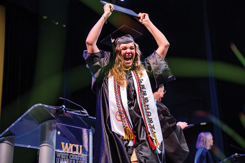 A student wearing graduation robes excitedly holds her diploma in the air