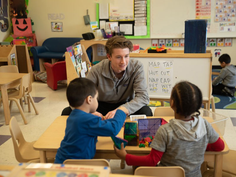 A student teacher sits at a small desk with two small children