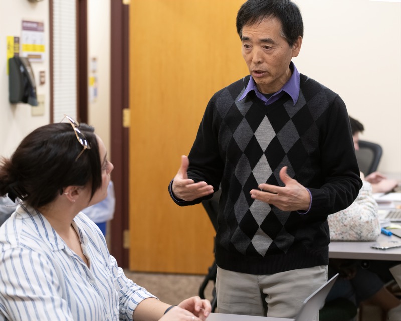 An Asian adult standing while speaking with a student who is sitting.
