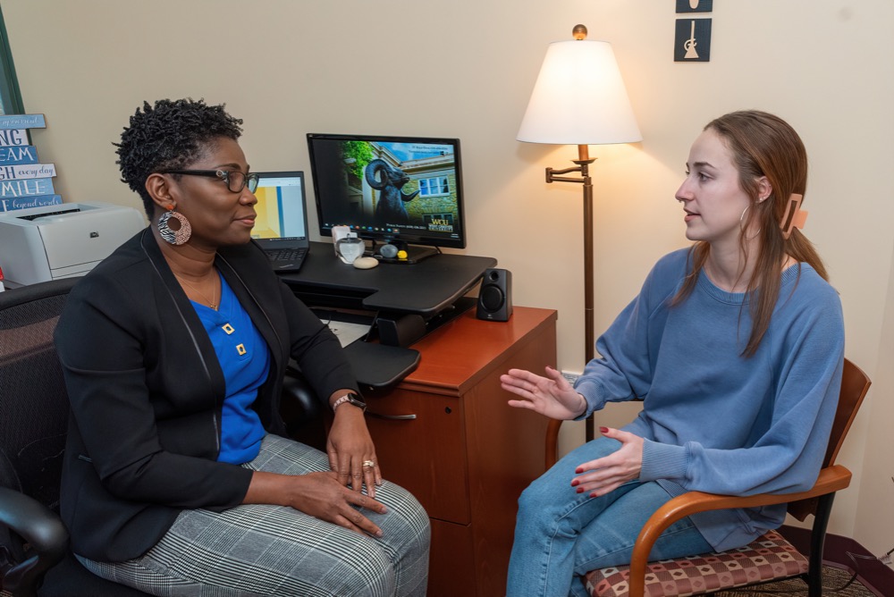 A woman of color speaking with a student. Both are sitting in an office environment