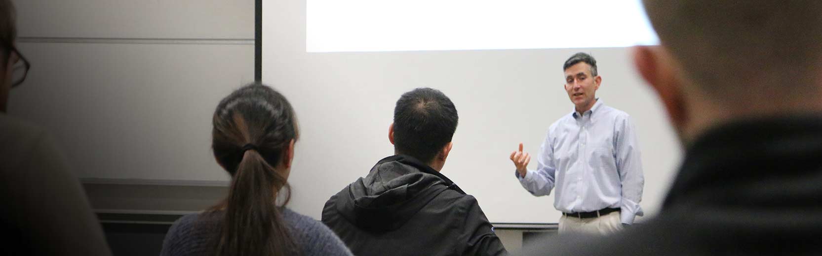 Instructor speaks at the front of a classroom, gesturing toward a projection screen while students sit and listen.
